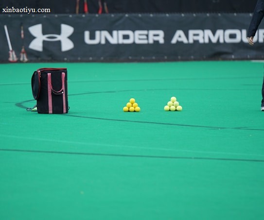 A sports field features a black and pink sports bag, two pyramids of yellow balls, and a portion of a person in dark athletic gear. The background displays a banner with the Under Armour logo.