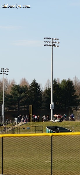 A sports field with a group of people engaging in athletic activities in the background. There is sports equipment like a soccer goal and a scoreboard visible. A chain-link fence runs across the foreground, and a statue or sculpture resembling an eagle is also seen. Trees and tall floodlights are positioned behind the field.