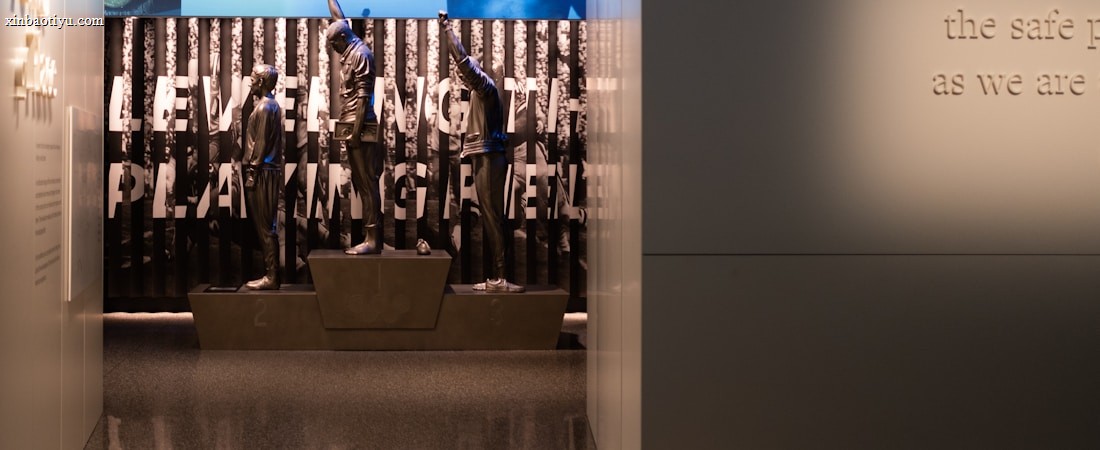 A dimly lit display features statues of athletes standing on a podium. They are positioned in front of a backdrop with the words 'LEVELING THE PLAYING FIELD' in bold letters. On the wall to the right, a softly lit quote is partially visible. The overall ambiance suggests a museum or exhibition setting.