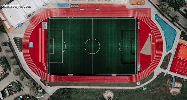 An aerial view of a large sports complex featuring a central green football field surrounded by a red running track. Adjacent to the track, there is a parking area, buildings, and pathways. Nearby, there are additional sports courts in different colors, including a blue swimming pool and orange basketball courts.