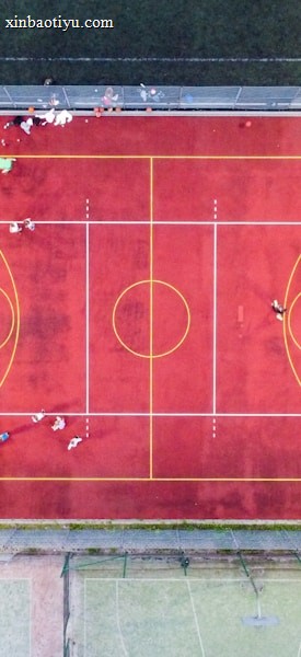 An aerial view of a sports court with a vibrant red surface and yellow lines marking different sections for basketball and soccer. Several people are present on the court, some engaged in activities, while others are standing along the perimeter. The court is enclosed by a metal fence and surrounded by grass on all sides.