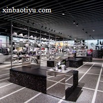 A modern sneaker store with sleek black shelving displaying various athletic shoes and sports apparel. Central seating areas and spotlights provide a contemporary retail ambiance. The signage prominently features a famous sportswear slogan in the background.