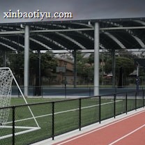 An outdoor sports facility featuring a running track and a soccer goal. The track is red, surrounded by a black metal fence, and the field is covered with artificial green turf. In the background, a basketball court is visible under a covered roof with metal supports. The sky is overcast, adding a moody atmosphere.